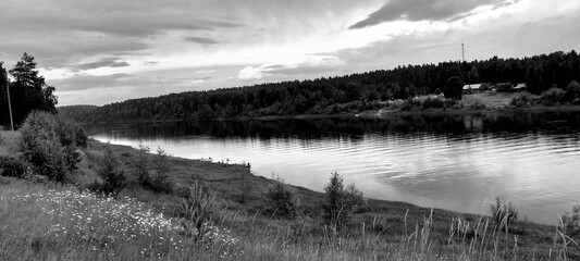 A serene black and white landscape shows a calm river reflecting the cloudy sky, bordered by grassy...