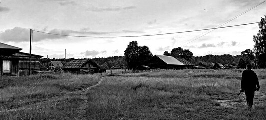 A black-and-white image shows a solitary figure walking through a grassy field. In the distance,...