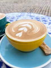 Yellow coffee cup with latte art sits on blue patterned table, showcasing intricate design and inviting atmosphere for relaxation and enjoyment during summer vacation