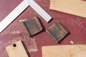 Woodworking elements arranged on a reddish surface. Two dark grooved blocks, two screws, a carpenter square, and light wood pieces