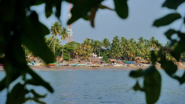Fishing village with boats and palm trees seen through leaves, calm and tropical
