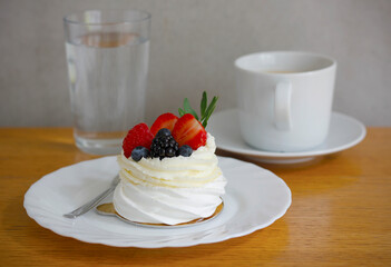 Small pavlova cake with whipped cream and fresh berries served on white porcelain plate with a glass of water and coffee cup on wooden table, minimal and natural light food styling
