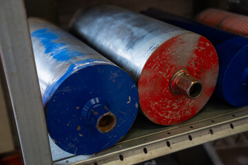 Industrial printing press rollers stored on metal shelf with worn paint and threaded shafts used in...