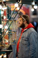 Vertical street photo. Mature white man with beard, carnival deer horns, blue denim jacket, red hoodie chooses gift on market stall with jewelry. Merry Christmas city holiday fair. Happy New Year 2026