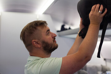Horizontal photo. White fat man with stubble, green t-shirt puts black backpack on luggage rack, baggage shelve in train close up. Concept of travel, tourism, arrival, summer vacation, road trip