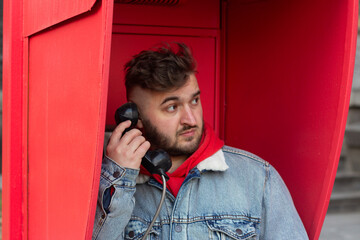 Horizontal street photo. Mature fat white man in jeans jacket, red hoodie calls from stationary payphone in retro telephone city booth close up. Concept of public phone, call, trip, travel, tourism, 