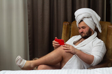 Horizontal photo. Young adult fat man with beard, white towel on head, robe sits on chair in hotel...