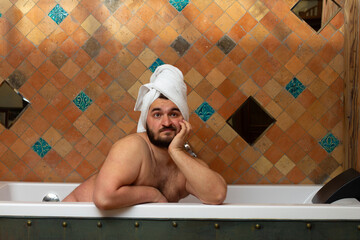 Horizontal photo. Young adult naked man with white towel on head in bathtub in hotel bath room with...