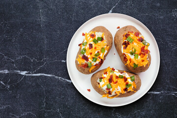 Loaded baked potatoes with cheese, sour cream, bacon and onions. Top view over a dark stone background.