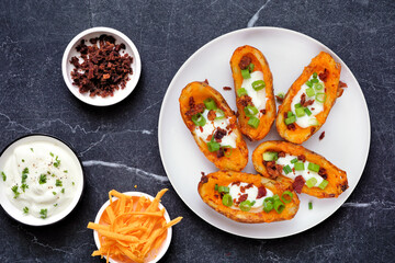 Loaded potato skins with cheese, sour cream, bacon and onions. Top view table scene over a dark stone background.