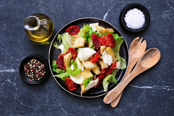 Italian salad with fresh mozzarella cheese, sun dried tomatoes, artichokes and lettuce. Top view table scene on a dark stone background.