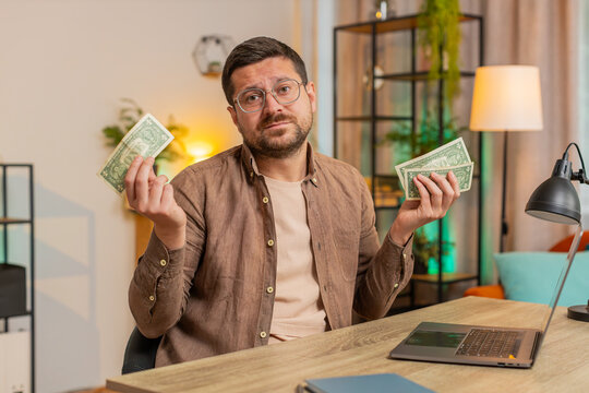 Caucasian man freelancer counting dollar cash insufficient amount of money. Financial crisis. Bankruptcy. Poverty and destitution. Upset businessman with laptop computer sitting at home office table. - Powered by Adobe