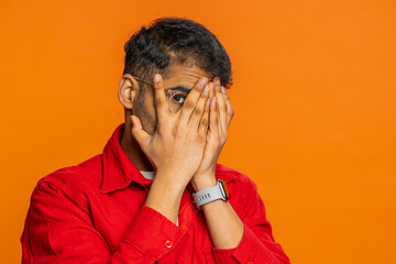 Nosy curious Indian man closing eyes with hand and spying through fingers, hiding and peeping,...