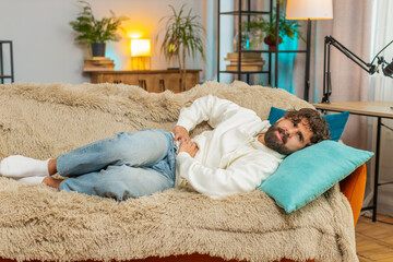 Young Indian man lying on sofa feeling sudden strong abdominal stomach ache, gastritis problem....