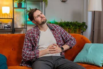 Caucasian young man sits on sofa feeling sudden strong abdominal stomach ache, gastritis problem....