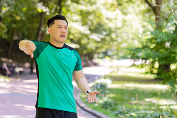 Dislike. Upset sportsman guy showing thumbs down sign gesture, expressing discontent disapproval dissatisfied. Displeased young Japanese man athlete looking at camera standing on footpath in park.