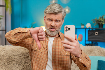 Mature man sitting on home sofa holding smartphone, showing thumbs down dislike gesture....