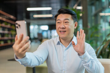 Portrait of smiling young businessman holding smartphone waving hand sign hello greeting...