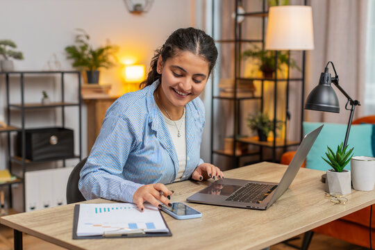 Indian freelancer accountant working with financial graph documents, calculate taxes for charts and reports at home office desk. Woman girl working on laptop phone planning expenses budget. Lifestyle