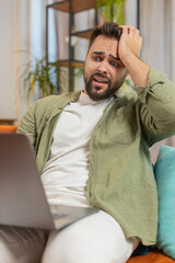 Upset young Caucasian young man sitting on sofa at home using laptop having anxiety and stress....