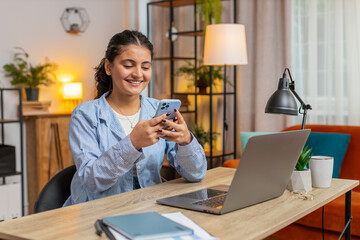 Happy young Indian woman sitting at table using smartphone smiling at home apartment. Girl freelancer texting share messages on mobile phone social media applications online watching movie. Lifestyle.