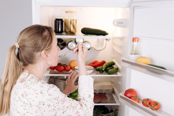 Woman examining fresh vegetables and dairy products in a refrigerator, showcasing healthy eating...