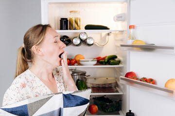 Woman yawning while holding a pillow, stands in front of an open refrigerator filled with fresh...