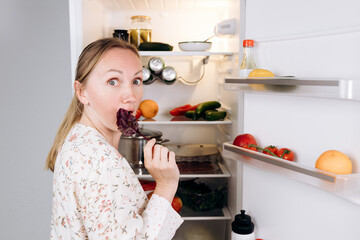 Woman with long hair enjoying fresh salad greens from refrigerator, surrounded by colorful...