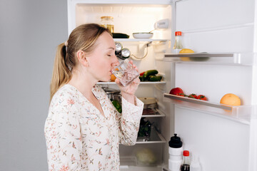 Woman enjoying refreshing glass of water in morning, standing in front of open refrigerator filled...