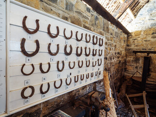 Fototapeta premium White wooden stand on a old stone wall with all types of rusty horse shoe. Blacksmith craft and skill. Farm and castle building. Vintage items show.