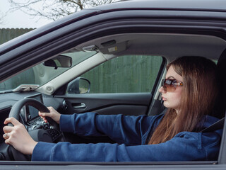 Naklejka premium A young inexperienced woman is driving a car with her sunglasses on. She is wearing a blue hoodie. High cost of insurance for young drivers concept. Travel and tourism concept.