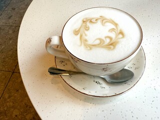 A cup of white coffee with a spoon on a saucer, sitting on a white table. Concept of: Simple coffee pleasure.