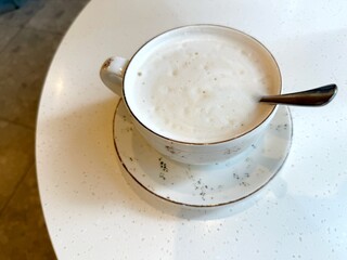 A cup of cappuccino with latte art sits on a saucer with a spoon on a white table. Concept of: Coffee break delight.