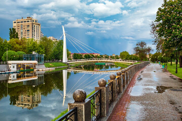 Cityscape with view of Kharkiv river, pedestrian iron bridge and buildings. Embankment with stone fence. Cloudy summer day, after rain