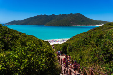 Pontal do Atalaia, Brazil, March 5, 2025. A walkway to a paradisiacal Brazilian beach, with turquoise waters, surrounded by green mountains, with tourists enjoying the sun and lush nature.