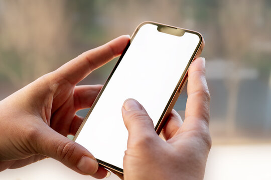 Manos sosteniendo un smartphone moderno con pantalla blanca, listo para insertar contenido. Fondo desenfocado, luz natural, enfoque en el dispositivo.