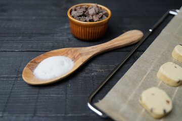 Top view of cut cookie dough on baking paper over rack with sugar in spoon and chocolate chips in bowl on black wooden background