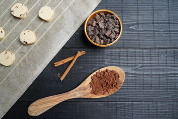 Top view of cookie dough on baking paper with wooden spoon of cacao, chocolate chips in brown bowl and cinnamon sticks on black wood