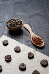 Top view of cut cookie dough on baking paper with cacap in spoon and chocolate chips in bowl on black wooden background