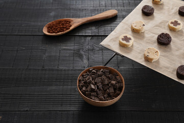 Front view of cut cookie dough on baking paper with wooden spoon of cacao, chocolate chips in brown...
