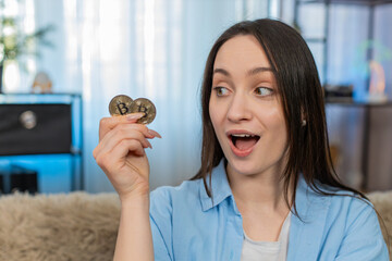 Young happy woman at home excitedly holding bitcoin BTC coin surprised by cryptocurrency profit,...