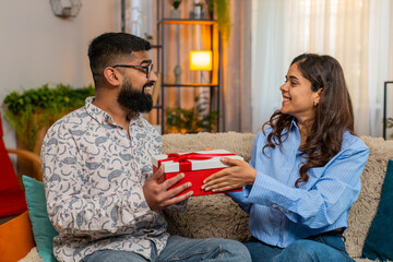 Indian woman and sitting on home sofa, presenting man gift box with joyful surprise and emotional...