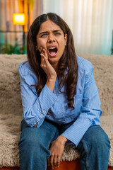 Indian young woman presses cheek in pain from sudden toothache while sitting on home sofa,...