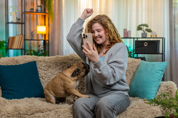 Young Caucasian redhead woman reads great news on smartphone while on home sofa, smiling with joy...