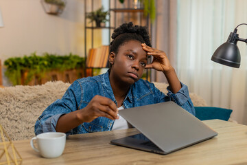 African American young woman closes laptop with visible stress after a hard day of online learning while sitting at table on home sofa. Black girl holds head, overwhelmed by digital study fatigue.