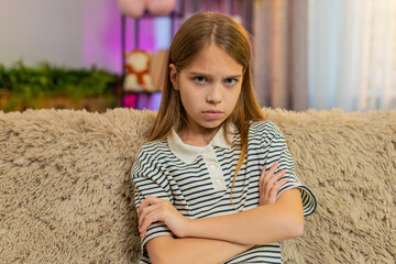 Young child girl crosses arms angrily sitting at home on sofa, showing clear signs of irritation...