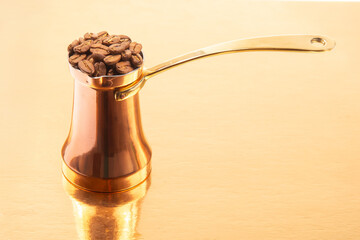 A polished copper cezve with a brass handle alongside a pile of coffee beans on a golden surface,...