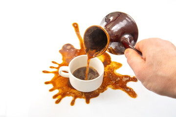 A hand pours coffee from a traditional pot into a white cup, spilling over with a large splash on a...