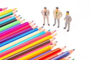 An assortment of vivid colored pencils lies beside three small business figurines on a clean white...