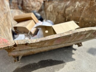 An old rusty dumpster sitting on a bustling city street filled with urban life and debris. The dumpster resting on the pavement clearly shows numerous signs of urban decay and waste management issues.